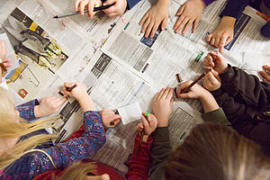 Kinderhände schreiben etwas auf Zettel über einer Zeitung.
