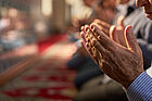 Gefaltete Hände im Bildvordergrund, dahinte betende Menschen in einer Moschee