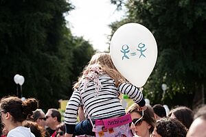 Kind sitzt bei einer Demonstation auf der Schulter eines Elternteils und hält einen Luftballon