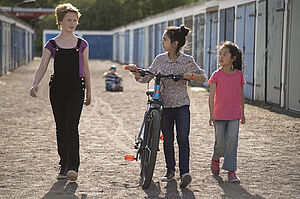 Drei Mädchen laufen nebeneinander her. Eins der Mädchen schiebt ein Fahrrad.