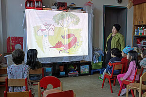 Kinder im Kita-Alter sitzen auf Stühlen und schauen auf eine improvisierte Kino-Leinwand. Eine Frau mit Kopftuch steht daneben.
