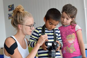 Eine junge Frau zeigt einem Jungen und einem Mädchen eine digitale Spiegelreflex-Kamera.