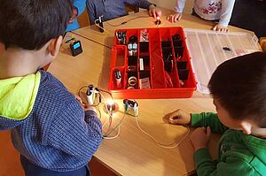 Zwei Kinder sitzen am Tisch, vor ihnen eine rote Box. Sie forschen zum Thema ‚Strom und Energie‘