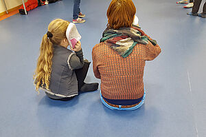 Ein Mädchen und eine Frau sitzen auf dem Boden und haben jeweils einen Schuh am Ohr